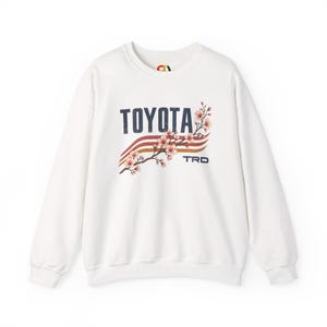 May include: A white crewneck sweatshirt featuring a graphic print on the chest. The design includes the word TOYOTA in dark blue, three horizontal wavy stripes in red and orange, a branch of pink cherry blossoms, and the letters TRD in blue.