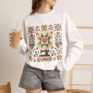 May include: A white long-sleeve sweatshirt featuring a graphic print of a vintage sewing machine, colorful spools of thread, heart shapes, and floral quilt-inspired patterns in shades of red, green, yellow, and blue on the front.