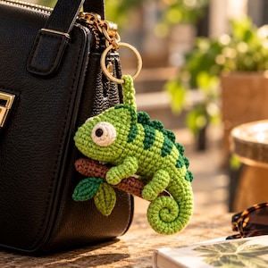 Op de afbeelding: Een gehaakte kameleon sleutelhanger in tinten licht- en donkergroen, met een groot wit oog en een zwarte pupil. De kameleon zit op een kleine bruine gehaakte tak met groene blaadjes, bevestigd aan een goudkleurige metalen sleutelring.