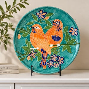 Può includere: Un piatto decorativo rotondo in ceramica con un vivace sfondo verde acqua e due uccelli arancioni appollaiati su un ramo. Il design include foglie verdi e piccoli fiori blu e arancioni. Il piatto è esposto su un supporto in metallo nero.