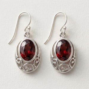 May include: A pair of silver drop earrings featuring oval-cut deep red garnet gemstones. Each stone is set within a polished silver frame decorated with intricate swirling metalwork patterns and attached to a curved silver ear wire hook.