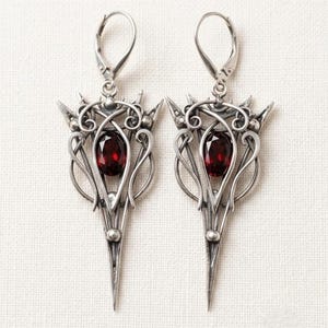May include: A pair of silver-toned dangle earrings featuring an intricate, pointed metal frame with swirling wire details. Each earring is centered with a single, oval-cut deep red gemstone and is finished with a secure lever-back closure.