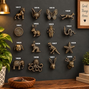 Può includere: Una collezione di sedici pomelli per mobili in metallo color bronzo con varie forme di animali e natura, ciascuno numerato da 1017 a 1032. I disegni includono un leone, una volpe, una farfalla, un coniglio, un serpente, un cane, un dinosauro, una stella marina, un elefante, un polpo e una testa di cervo.