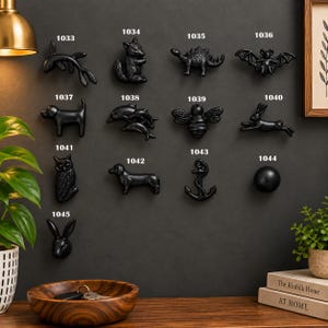May include: A collection of thirteen black decorative knobs arranged on a wall, each labelled with a number from 1033 to 1045. The shapes include a fish, squirrel, dinosaur, bat, dog, dolphins, bee, rabbit, owl, dachshund, anchor, sphere, and rabbit head.