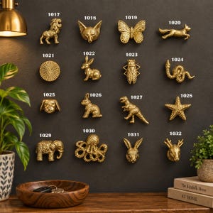Könnte beinhalten: Eine Sammlung von sechzehn goldfarbenen Metall-Möbelknöpfen, die in einem Raster an einer dunklen Wand angeordnet sind. Jeder Knopf zeigt ein einzigartiges Tier oder eine Form, darunter Löwe, Fuchs, Schmetterling, Hase, Schlange, Hund, Dinosaurier, Seestern, Elefant, Krake und Hirsch, nummeriert von 1017 bis 1032.
