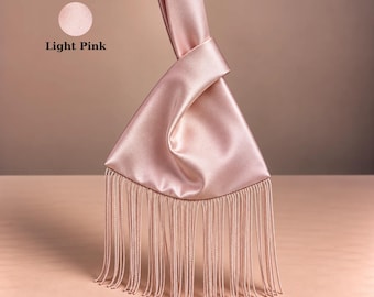May include: A light pink satin wristlet handbag featuring a knotted handle design and a long, flowing fringe trim along the bottom edge. The bag has a smooth, reflective surface and includes a circular icon with the text Light Pink.