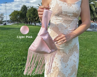 Peut inclure: Un sac à main poignet en satin rose clair doté d'une anse nouée et de longues franges fluides à la base. Le tissu présente un éclat lisse et réfléchissant, et le texte Light Pink est imprimé à côté d'un échantillon de couleur circulaire.