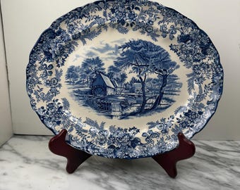 May include: An oval ceramic platter featuring a blue and white transferware pattern. The center depicts a rural landscape with a water mill and trees, while the scalloped rim is decorated with a dense floral and botanical motif.