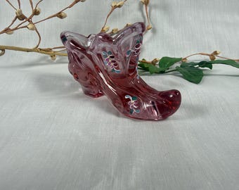 May include: A translucent pink glass shoe figurine featuring a molded, curved silhouette and delicate hand-painted floral accents in shades of teal, white, and purple along the exterior surface of the glass boot structure.
