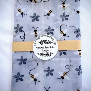 May include: A rectangular beeswax wrap featuring a light blue background with a repeating pattern of yellow and black bees, dotted flight paths, and dark blue flowers. A tan paper band holds a circular label reading Sourdoughsweetiebakery.com Natural Bees Wax Wraps.