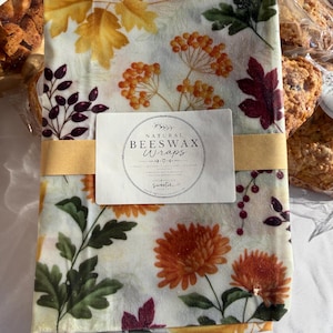 May include: A rectangular beeswax wrap featuring a cream background with a colorful autumn-themed print of orange chrysanthemums, yellow maple leaves, dark red berries, and green foliage, secured by a tan paper band with the text Natural Beeswax Wraps.