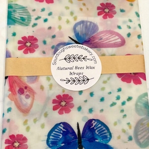May include: A rectangular beeswax wrap featuring a white background with a colorful pattern of pink flowers, teal dots, and blue, purple, and yellow butterflies. A tan paper band with a circular label reads Sourdoughsweetiebakery.com Natural Bees Wax Wraps.
