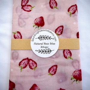 May include: A rectangular beeswax wrap featuring a light pink background with a repeating pattern of red strawberries and small white dots. A tan paper band wraps around the center with a circular label reading Sourdoughsweetiebakery.com Natural Bees Wax Wraps.