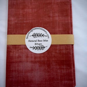 May include: A rectangular, folded piece of fabric with a textured, terracotta-red finish. A tan paper band wraps around the center, featuring a circular white label with the text "Sourdoughsweetiebakery.com Natural Bees Wax Wraps" and leaf icons.