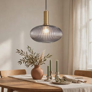 Peut inclure: Une suspension lumineuse composée d'un abat-jour en verre gris fumé strié, fixé à une monture métallique cylindrique de couleur laiton. La lampe est suspendue par un fin cordon noir et contient une ampoule allumée diffusant une lumière douce.
