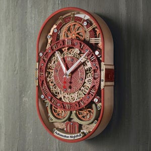 May include: An oval wall clock with a red frame and intricate internal gears in copper and gold tones. The clock face features Roman numerals, white hands, and a red second hand. A small plaque at the bottom displays the text Automaton Nightfall.