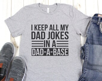 May include: A gray t-shirt with the text "I keep all my dad jokes in a dad-a-base" printed in black. The shirt is laid flat on a white wooden surface with a pair of gray sneakers and blue jeans.