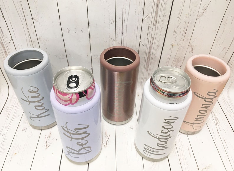 May include: Five personalized stainless steel can coolers. The coolers are in various colors, including gray, white, rose gold, and pink. Each cooler has a name engraved on it: Katie, Beth, Madison, and Amanda.