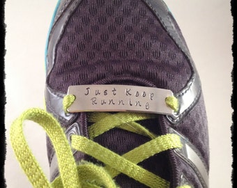 Könnte beinhalten: Nahaufnahme eines Laufschuhs mit einem lila Mesh-Obermaterial, silbernen Akzenten und leuchtend grünen Schnürsenkeln. Ein kleines Metallschild mit der Aufschrift "Just Keep Running" ist an den Schnürsenkeln befestigt. Der Schuh ist für sportliche Aktivitäten konzipiert.