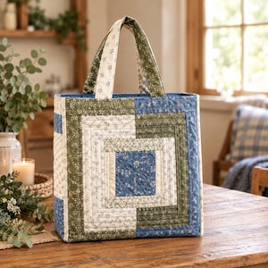 May include: A quilted tote bag featuring a log cabin block pattern in shades of blue, olive green, and cream. The fabric displays a small floral print throughout, with two matching fabric handles attached to the top of the rectangular bag.