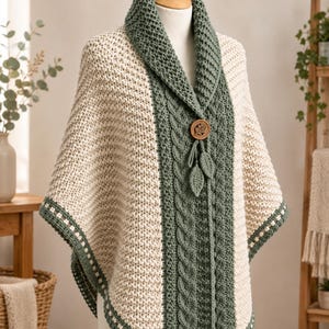May include: A cream and sage green knit poncho featuring a wide shawl collar, a central cable knit panel, and a single round wooden button closure. Two small crocheted leaf-shaped embellishments hang from the button, and the hem is finished with a decorative border.