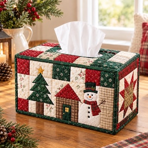 Op de afbeelding: Een rechthoekige, doorgestikte tissueboxhoes met een patchworkontwerp van rode, groene en crèmekleurige stoffen vierkanten. De voorkant toont een geappliqueerde kerstboom, een klein huisje en een sneeuwman met een zwarte hoge hoed.
