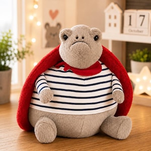 Puede incluir: Un peluche con forma de tortuga de cuerpo gris suave, que lleva una camiseta blanca con rayas horizontales azul marino y un pañuelo rojo brillante alrededor del cuello. La tortuga tiene un caparazón rojo grande, redondo y texturizado.