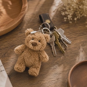 May include: A plush brown teddy bear keychain attached to a dark brown leather strap with a gold-toned metal snap button. The keychain features a silver-toned metal ring holding three assorted metal keys in silver and brass tones.