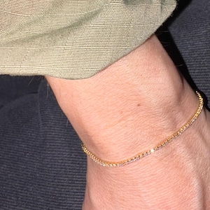 Può includere: Un delicato braccialetto d'oro con piccoli accenti scintillanti. Il braccialetto è indossato su un polso, con una manica di giacca mimetica visibile sullo sfondo. Il braccialetto ha una circonferenza di circa 18 cm.