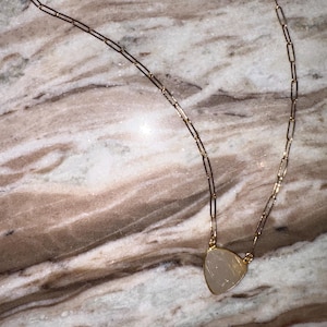 May include: A gold-colored necklace with a triangular pendant. The pendant is a light, translucent color. The chain is made of rectangular links and has a clasp closure.