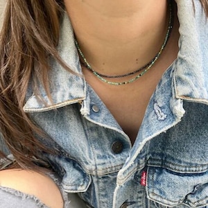 Peut inclure: Deux colliers de perles délicats, verts et gris foncé, reposent sur une veste en jean bleu clair. Les colliers semblent être faits de petites perles rondes, créant un effet superposé. La veste a un design classique.