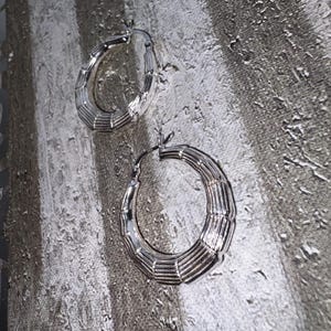 May include: A pair of silver hoop earrings with a ridged design. The earrings are circular and feature a series of horizontal lines, giving them a textured look. They are set against a textured, grey background.