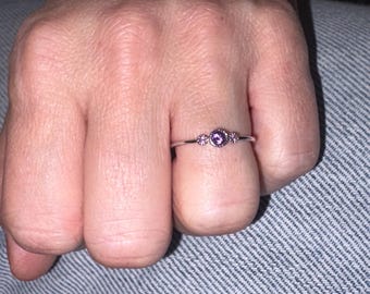 Puede incluir: Un anillo plateado con una gema púrpura central y dos piedras púrpuras más pequeñas a cada lado. El anillo está en un dedo, con un fondo neutro.