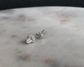 May include: A pair of silver earrings featuring a hamsa hand design, each hand is encrusted with small clear cubic zirconia stones.