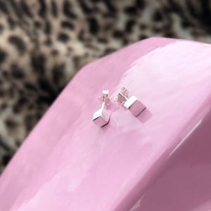 May include: A pair of silver square stud earrings on a pink surface.
