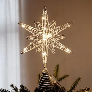 Puede incluir: Una estrella de alambre plateado para la punta del árbol con un diseño geométrico de varias capas y luces LED de color blanco cálido integradas. La estrella se asienta sobre una base de alambre en espiral en las ramas verdes.