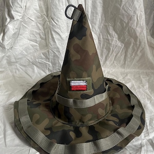 Puede incluir: Un sombrero cónico con un patrón de camuflaje en tonos verdes, marrones y negros. El sombrero incluye un ala ancha con correas de cincha integradas, una pequeña anilla en D negra en la punta y un parche rectangular con una bandera blanca y roja.