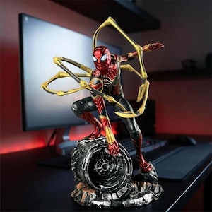 以下が含まれることがあります： 赤、黒、金のスーツを着たスパイダーマンのアクションフィギュア。背中から4本の機械的なスパイダーレッグが伸びている。フィギュアは、暗く風化した仕上げの工業的なスタイルの円形金属ベースの上にしゃがんでいる。