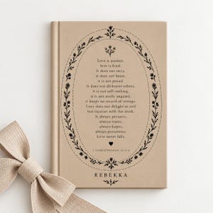 May include: A tan hardcover book featuring a black oval floral border enclosing the text of 1 Corinthians 13:4-8, ending with a small heart and the name REBEKKA. A tan fabric ribbon is tied in a bow at the bottom left corner of the book cover.