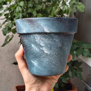 Peut inclure: Un pot de fleurs conique présentant une couleur de base bleu foncé et noire avec une finition marbrée rappelant une galaxie. La surface est décorée d'éclaboussures et de traînées de peinture blanche créant un aspect texturé et cosmique.