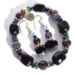 May include: A jewelry set featuring a bracelet and earrings. The bracelet and earrings are made with black, purple, and silver beads. The earrings have silver hooks. The jewelry set is on a white background.