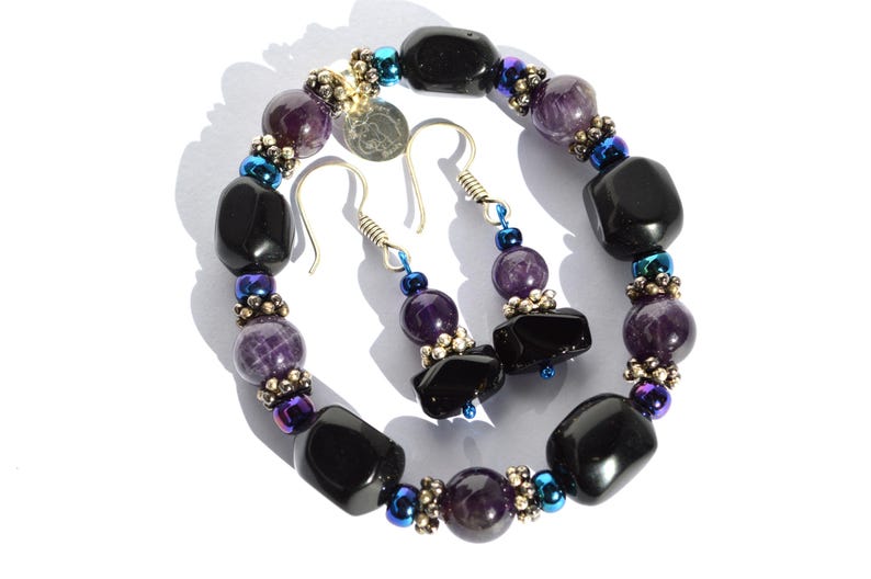 May include: A jewelry set featuring a bracelet and earrings. The bracelet and earrings are made with black, purple, and silver beads. The earrings have silver hooks. The jewelry set is on a white background.