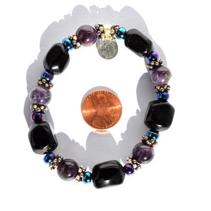 May include: A beaded bracelet featuring black and purple gemstones, interspersed with smaller iridescent beads. A small silver charm hangs from the bracelet. The bracelet is shown next to a penny for size reference.