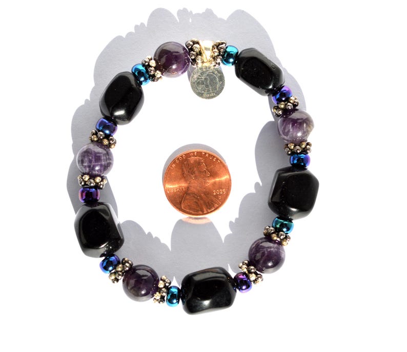 May include: A beaded bracelet featuring black and purple gemstones, interspersed with smaller iridescent beads. A small silver charm hangs from the bracelet. The bracelet is shown next to a penny for size reference.