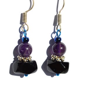 May include: A pair of handcrafted earrings featuring a silver-tone hook, blue accents, and a combination of beads. The earrings showcase a dark purple, round bead, a cluster of small silver beads, and a black, rectangular stone. The jewelry is set against a white background.