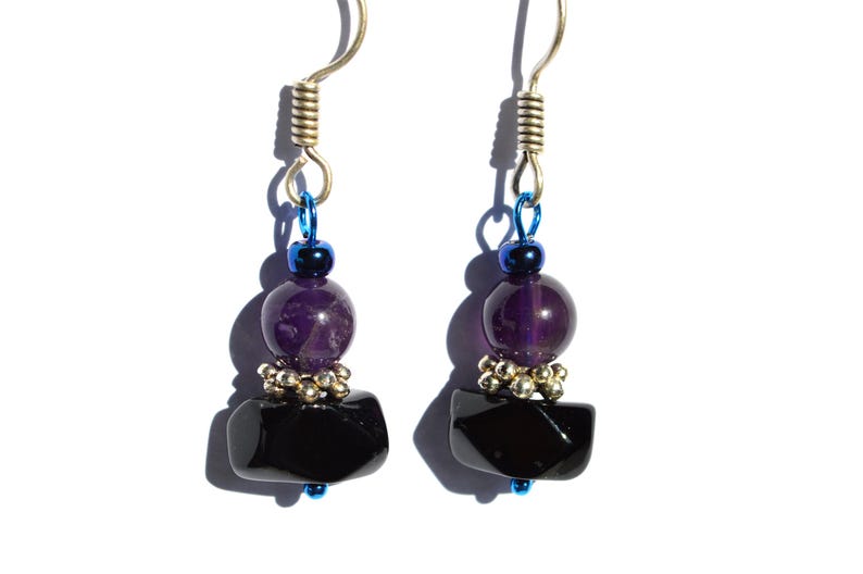 May include: A pair of handcrafted earrings featuring a silver-tone hook, blue accents, and a combination of beads. The earrings showcase a dark purple, round bead, a cluster of small silver beads, and a black, rectangular stone. The jewelry is set against a white background.