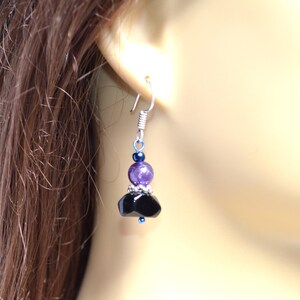 May include: A dangling earring with a silver hook and a cluster of beads. The earring features a small dark blue bead, a larger purple bead, and a black, faceted bead cluster. The earring is shown against a blurred background.