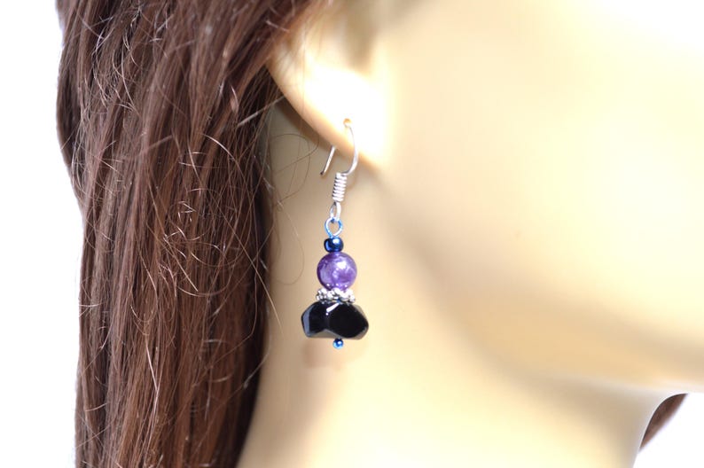 May include: A dangling earring with a silver hook and a cluster of beads. The earring features a small dark blue bead, a larger purple bead, and a black, faceted bead cluster. The earring is shown against a blurred background.