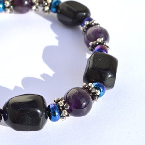 May include: A beaded bracelet featuring alternating black, purple, and blue beads. The bracelet has large, polished black beads interspersed with smaller purple and iridescent blue beads. Silver-colored spacers add detail to the design.