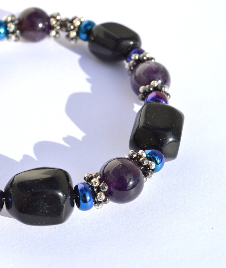 May include: A beaded bracelet featuring alternating black, purple, and blue beads. The bracelet has large, polished black beads interspersed with smaller purple and iridescent blue beads. Silver-colored spacers add detail to the design.